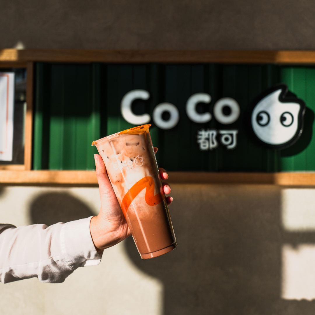 Someone holding a cup of CoCo bubble tea drink in front of a CoCo franchise shop. 